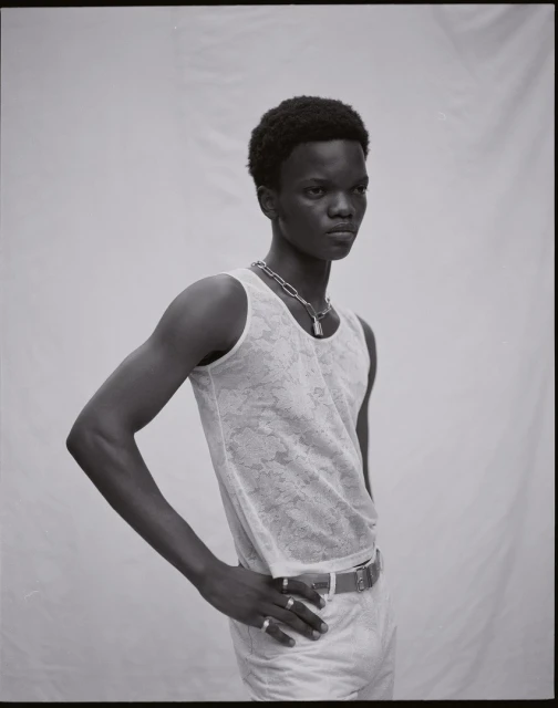 Portrait of a young adult Black male in a white tank top and light-colored pants, posing against a neutral background in a studio setting.