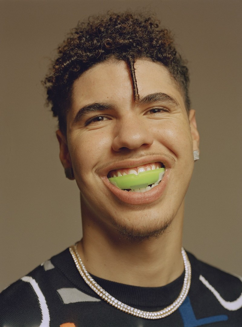 Close-up studio portrait of a smiling adult male wearing a mouthguard and jewelry, set against a neutral grey backdrop.