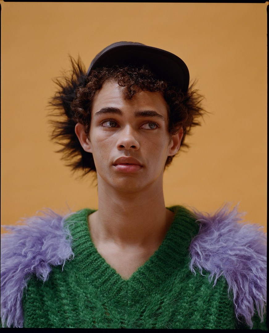 Portrait of a young adult male model with curly hair wearing a baseball cap and a green V-neck sweater with purple fur accents against an orange backdrop.
