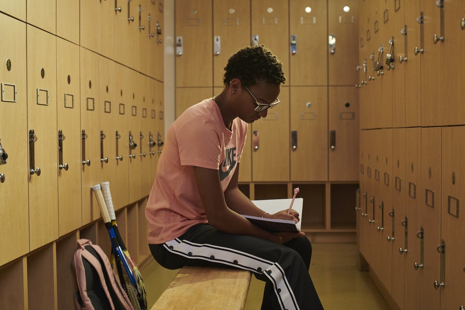 Young woman with glasses sits on a bench in a locker room, writing in a notebook next to tennis rackets and a pink backpack.