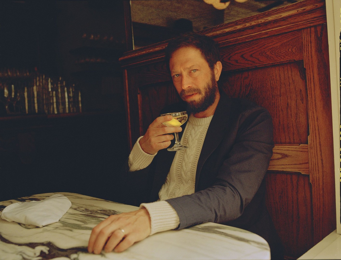 Portrait of an adult man with a beard, wearing a sweater and jacket, seated at a marble table while holding a cocktail glass indoors.