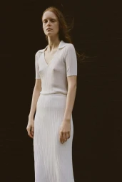 A woman with long reddish hair wearing a white ribbed polo shirt and skirt standing against a dark brick wall.