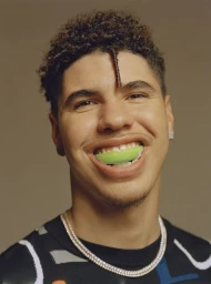 Portrait of a man with short curly hair wearing a dark patterned sweater and diamond chain necklace, smiling with a green mouthpiece.