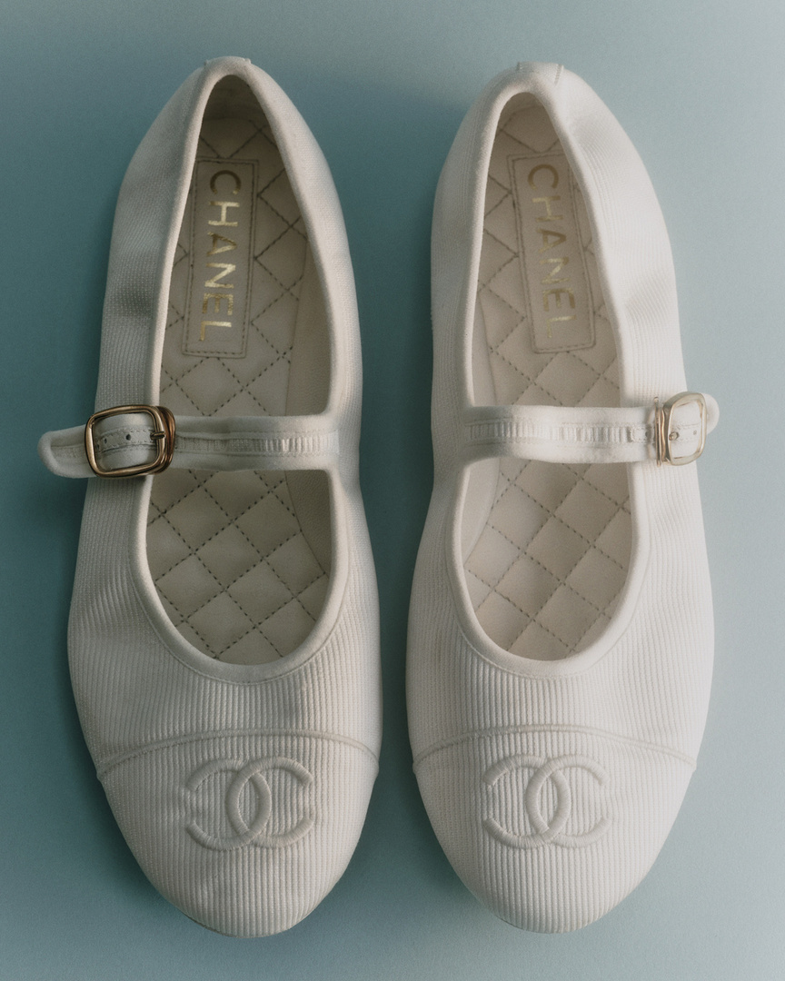 Pair of white Chanel Mary Jane ballet flats. Elegant footwear with gold buckle, classic design, displayed against a light blue background.