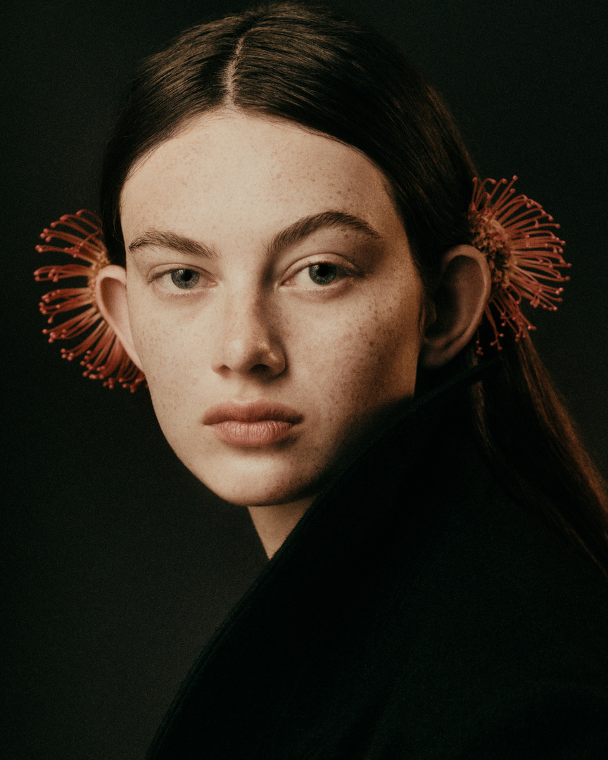 Portrait of a young woman with flowers behind her ears, wearing a dark coat against a dark grey background in a studio setting.