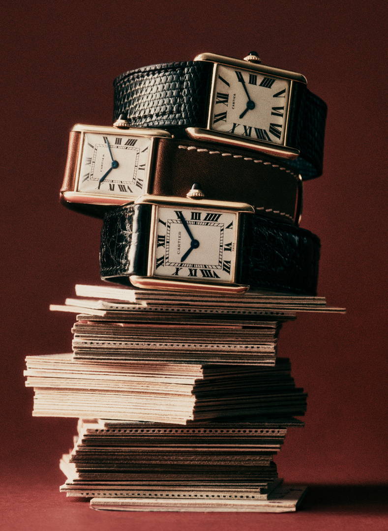 Still life studio shot of three Cartier watches stacked on top of each other, placed on a stack of postcards against a reddish-brown backdrop.