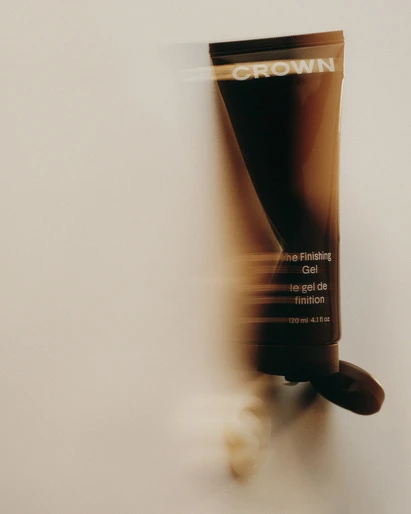 Crown The Finishing Gel product shot. Brown and black tube with brand name and product info on light beige background with motion blur.