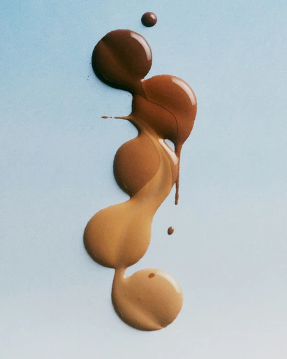 Several shades of foundation makeup in a vertical gradient from dark to light on a soft blue background. Beauty and cosmetic product.