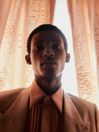 Portrait of a young adult Black male wearing a tan suit with a collared shirt, against a floral-patterned curtain backdrop in soft lighting.