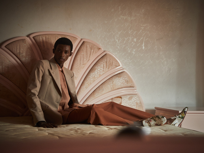 Person in beige suit reclines on a peach colored bed with a seashell-shaped headboard in an indoor setting with neutral color palette.