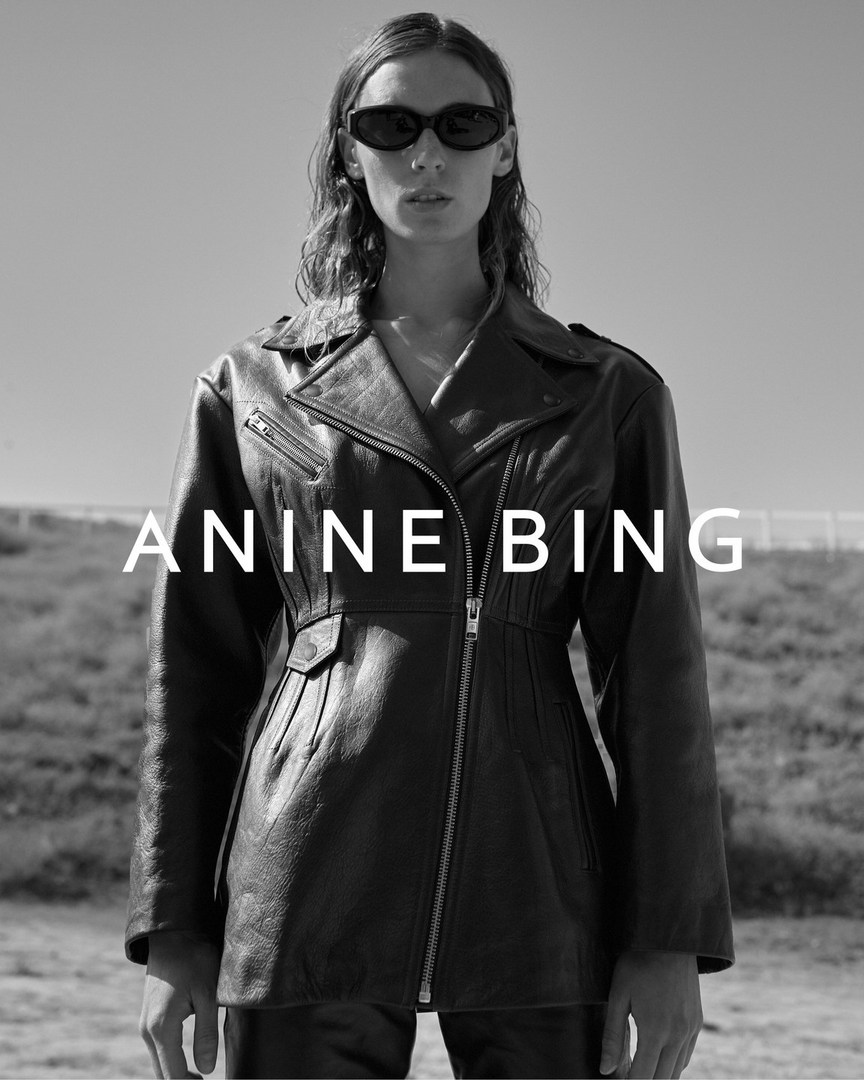 An adult female fashion model poses in a black leather jacket and sunglasses outdoors against a blurred blue sky background.