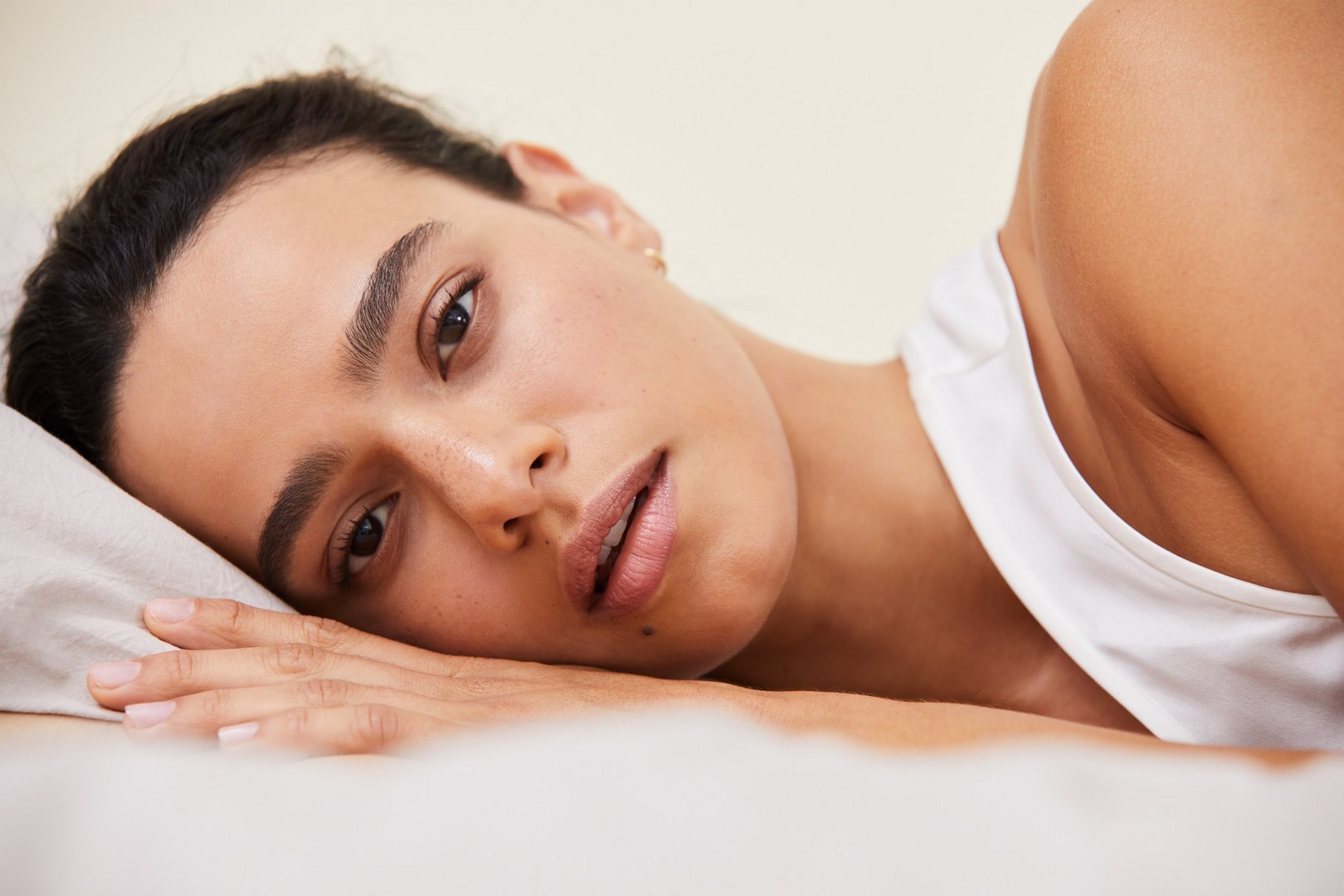 Close-up of an adult woman lying in bed with her head on a pillow, looking at the camera in a brightly lit indoor setting.