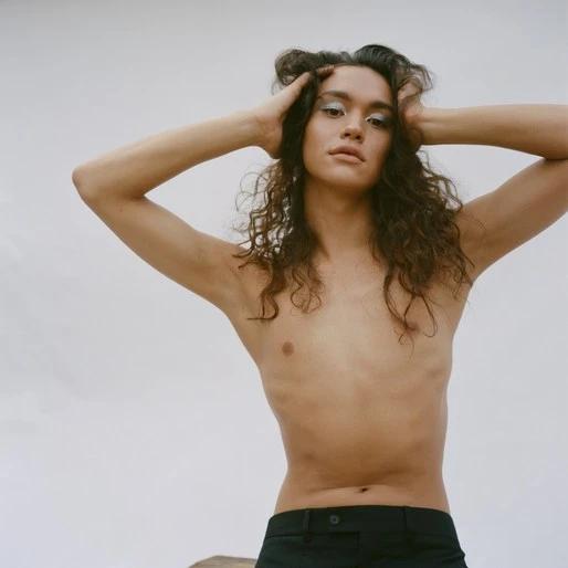 Androgynous person with long curly hair poses shirtless in a studio with hands on head and metallic eyeshadow on eyelids.