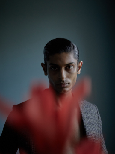 Portrait of a handsome adult male with slicked-back hair, wearing a blazer, with red bokeh in the foreground against a teal backdrop.