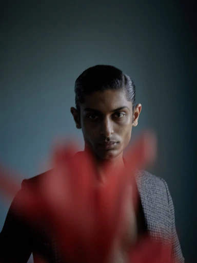 Portrait of a handsome adult male with slicked-back hair, wearing a blazer, with red bokeh in the foreground against a teal backdrop.
