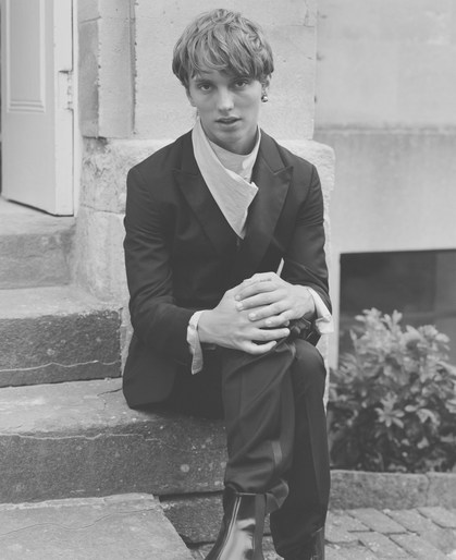 Monochrome portrait of a young adult male with tousled hair, seated on stone steps wearing a suit jacket and shiny boots outdoors.