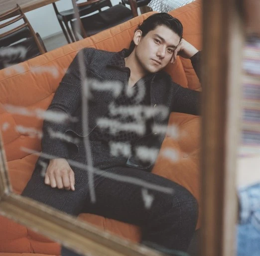 Portrait of an adult male model with dark hair reclining on an orange couch, as seen through a vintage picture frame.