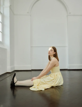 A woman in a yellow dress sits on a dark floor in a bright room with white arched walls and tall windows.