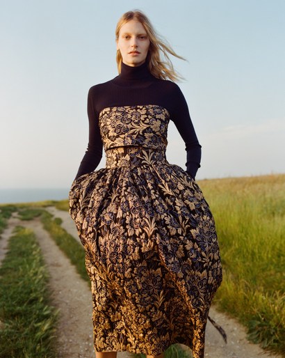 Woman in floral dress poses on grassy path under blue sky. Full-length fashion shot with natural light in a field.