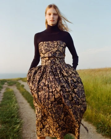 Woman in floral dress poses on grassy path under blue sky. Full-length fashion shot with natural light in a field.