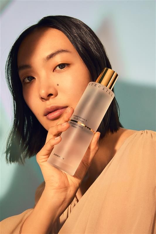 An Asian adult female is seen in a medium shot indoors, holding a bottle of skincare product while modelling and posing for a beauty advertisement.