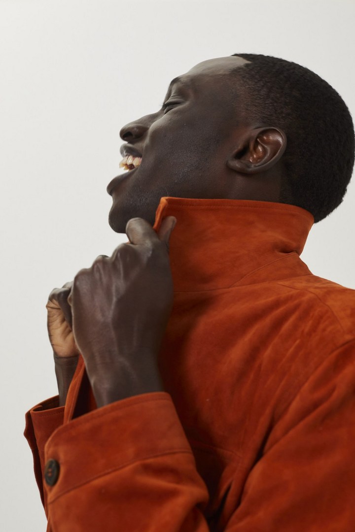 Profile shot of a Black adult male laughing, wearing an orange suede jacket with a raised collar against a bright white background.