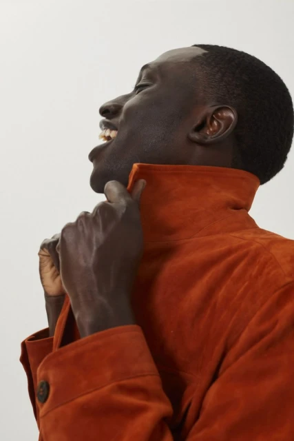 Profile shot of a Black adult male laughing, wearing an orange suede jacket with a raised collar against a bright white background.