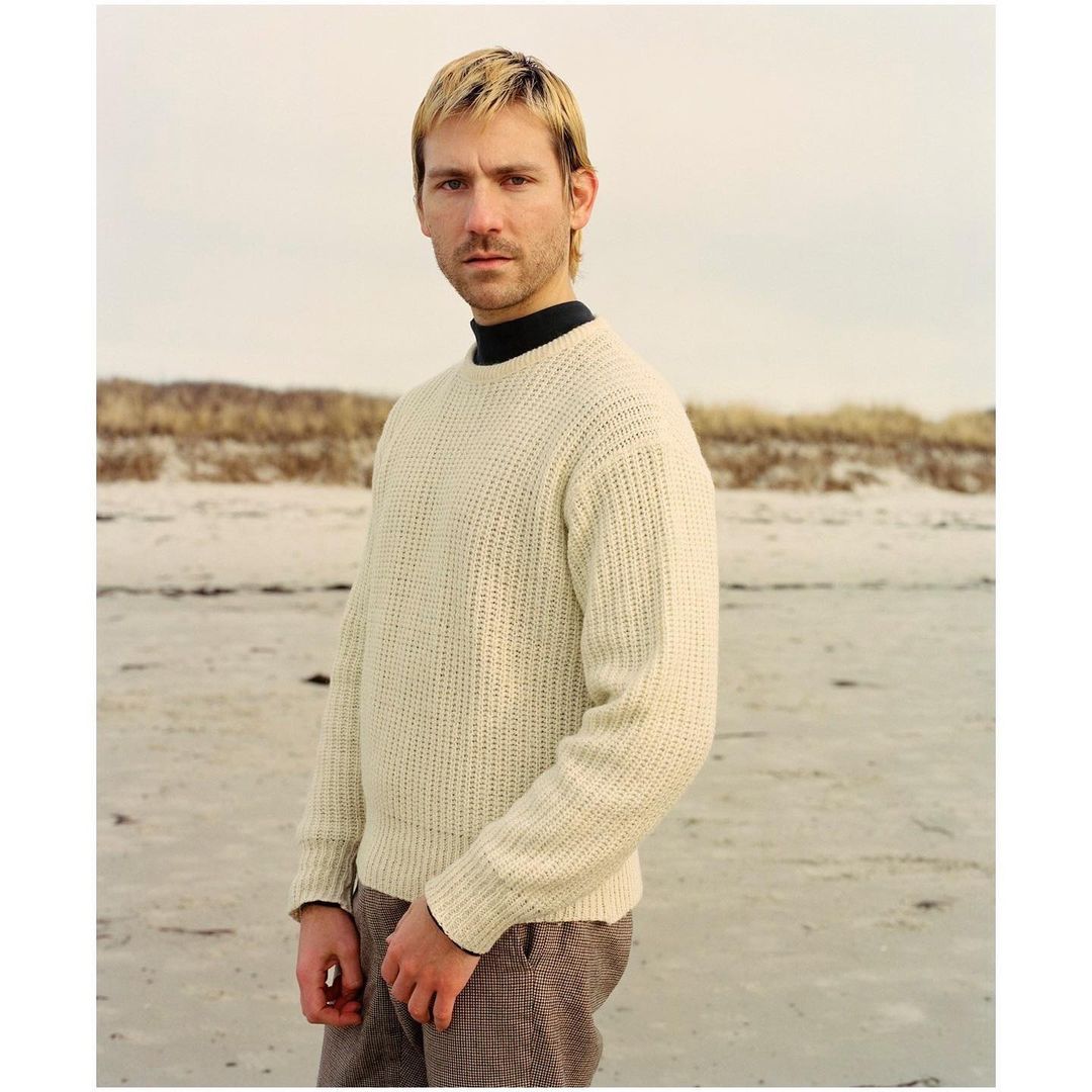 Man in cream sweater stands on sandy beach under overcast sky, portrait.