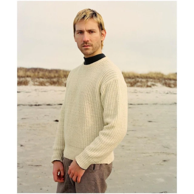 Man in cream sweater stands on sandy beach under overcast sky, portrait.