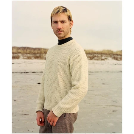 Man in cream sweater stands on sandy beach under overcast sky, portrait.