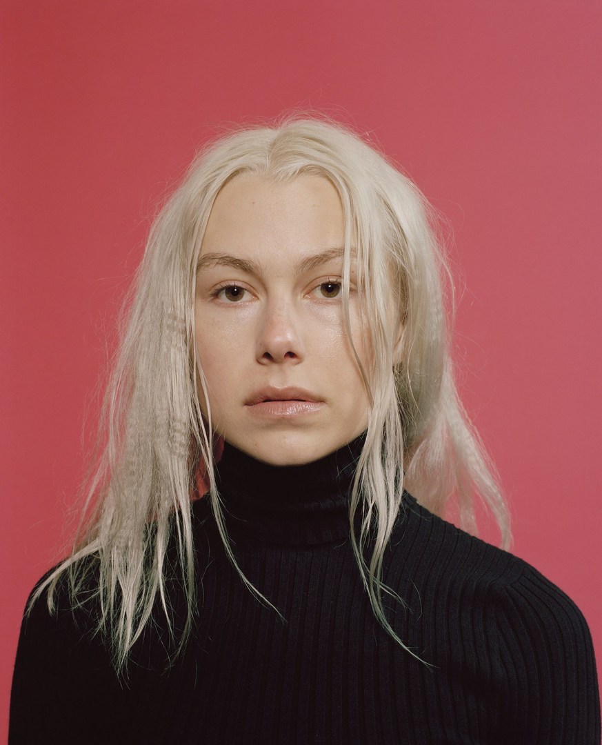 Close-up portrait of a young adult woman with fair skin, light blonde crimped hair, and a black turtleneck against a pink background.