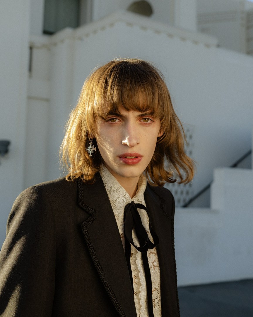 Portrait of a person with blond hair wearing a black jacket and a lace shirt posing outdoors against a white building backdrop.
