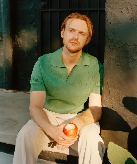 A fair-skinned adult male with light brown hair sits outdoors in natural light, holding a red apple, wearing a green shirt and tan pants.