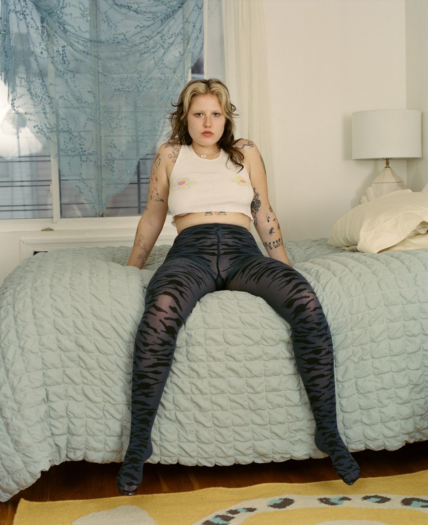 Adult female poses on a blue quilt bed with tattoo arm details, wearing a white crop top and tiger stripe leggings in a bedroom.