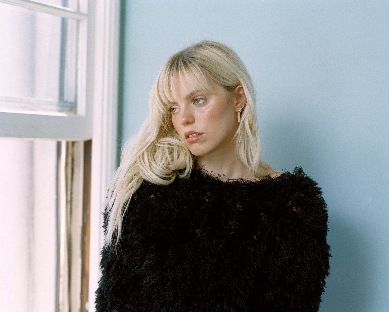 Blonde woman with bangs wearing a textured black top standing by a window against a blue wall