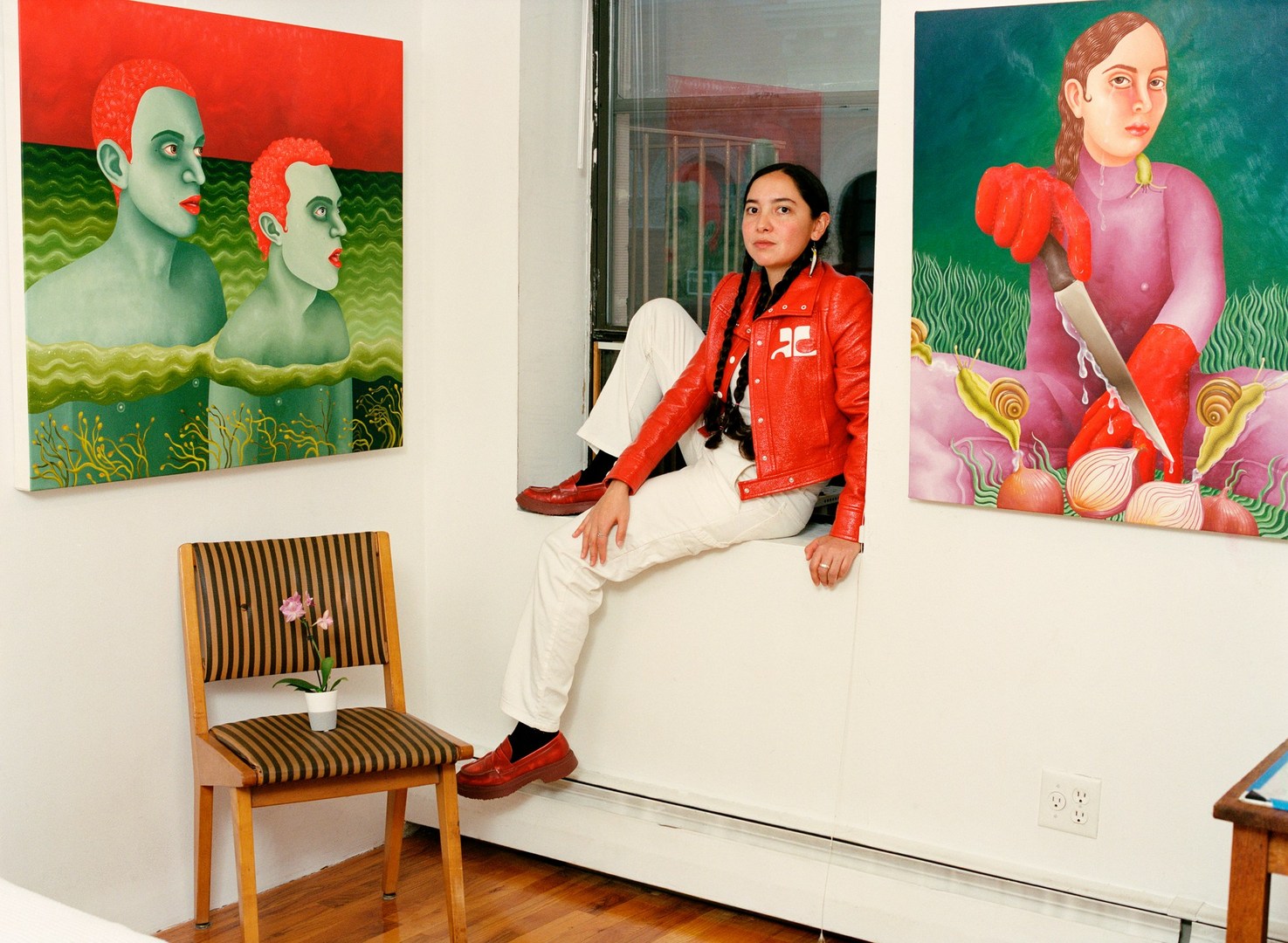 Adult female in red jacket and white pants poses seated against a white wall with artwork, chair, and hardwood flooring.