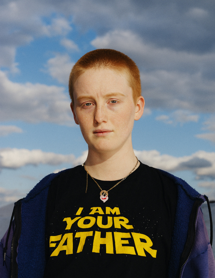 Portrait of a young adult woman with red hair and a black T-shirt with yellow text, standing outdoors against a cloudy sky.