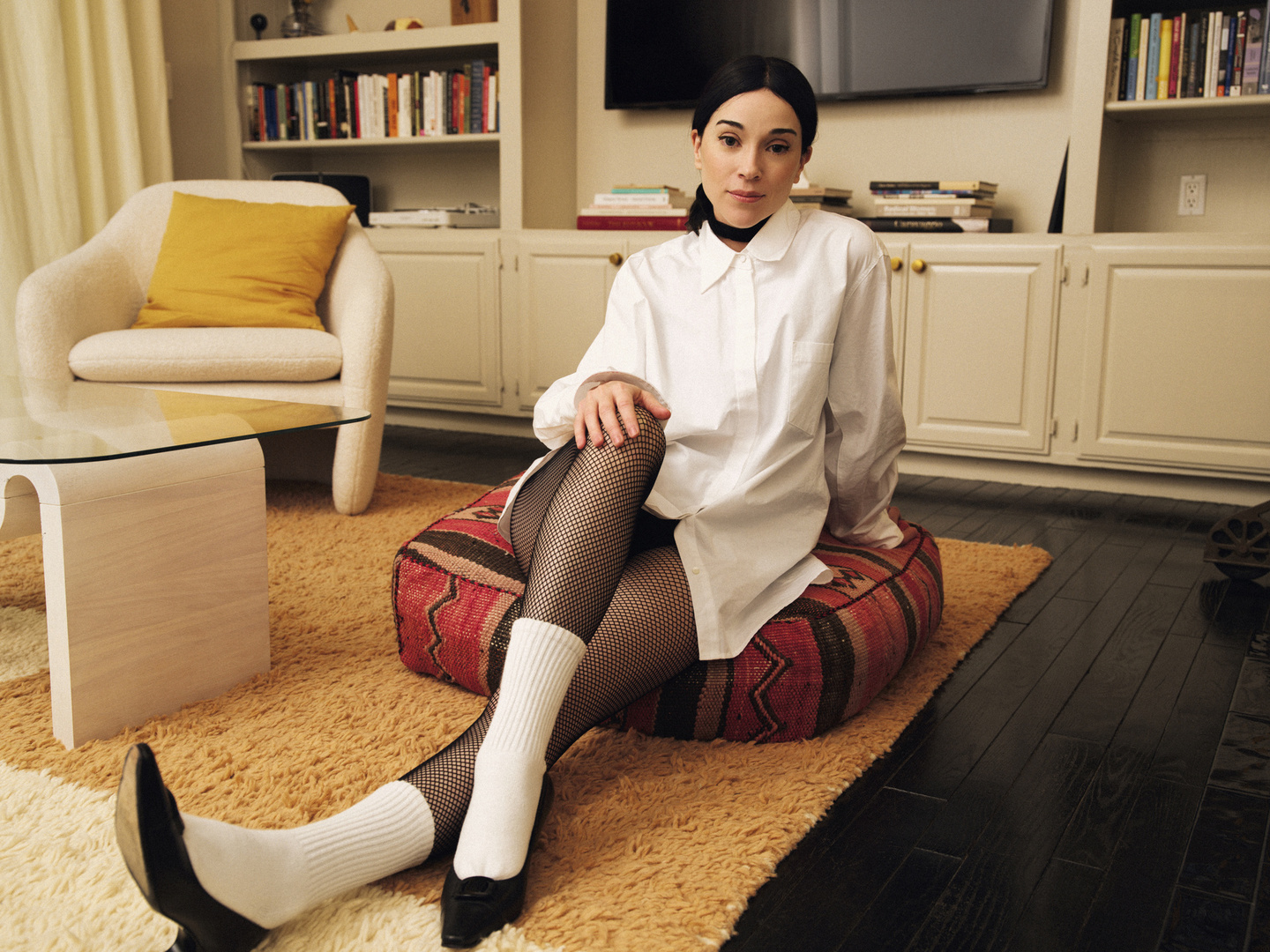 Woman in a white shirt, fishnet tights, and black heels poses on a patterned cushion, with a glass table and bookshelves in the background.