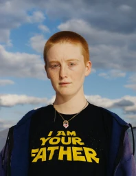 Portrait of a person with short red hair wearing a graphic t-shirt and necklace against a cloudy sky.