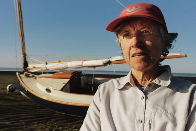 An elderly woman with short gray hair wears a red cap and stands in front of a blue and white sailboat on a beach under a clear sky.