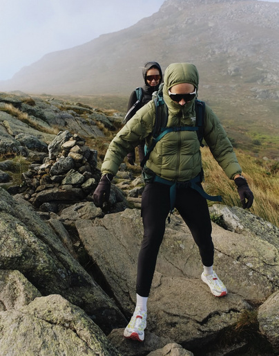 Two hikers traverse a rocky mountain landscape under overcast skies, clad in jackets and sunglasses, with a foggy peak in the background.