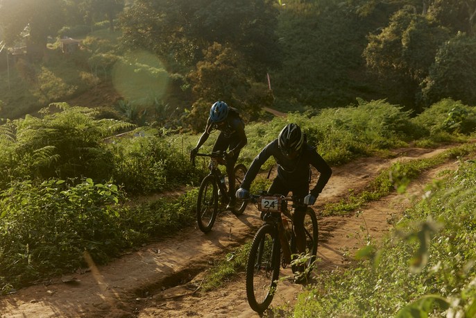 Two cyclists ride mountain bikes on a dirt path through a lush green landscape, wearing helmets and athletic gear in bright sunlight.