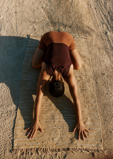 Woman in yoga pose on woven rug outdoors in sun. Brown shorts and tank top. Hands flat on mat, head down. Beige sand, sunny day.