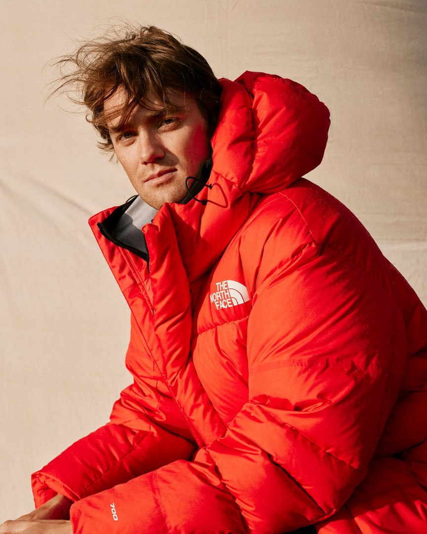 Portrait of a male with light hair wearing a red puffer coat by The North Face, shot with soft lighting against a neutral background.