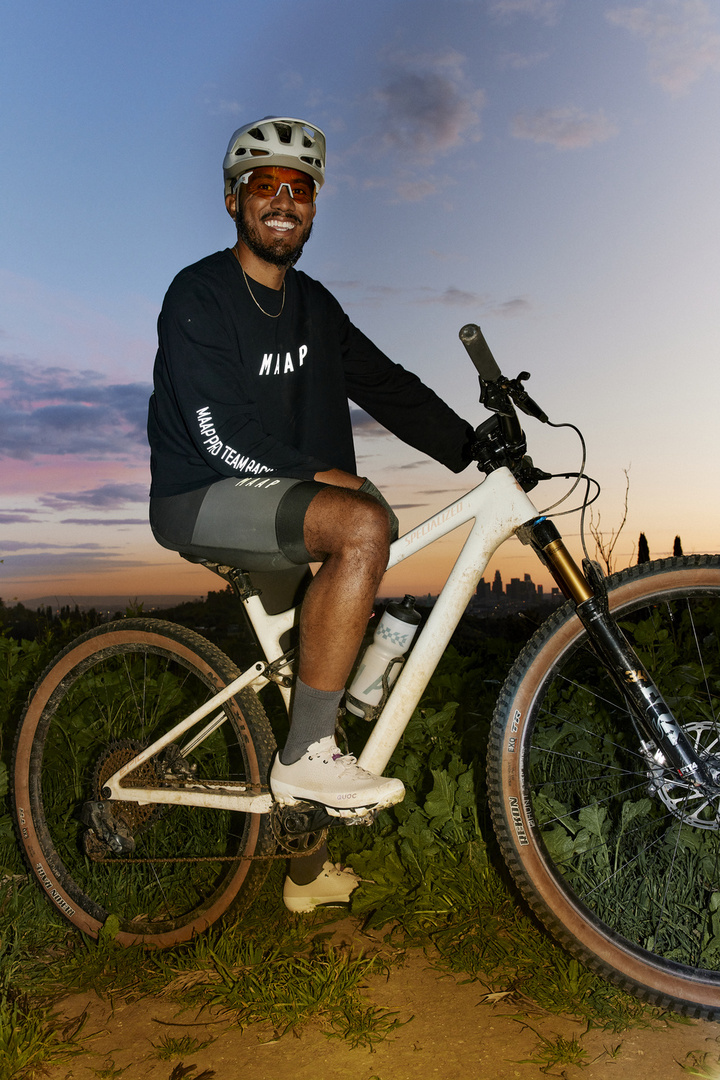 A cyclist pauses on a trail, seated on a mountain bike at dusk; a skyline is barely visible on the horizon beneath clouds and blue sky.