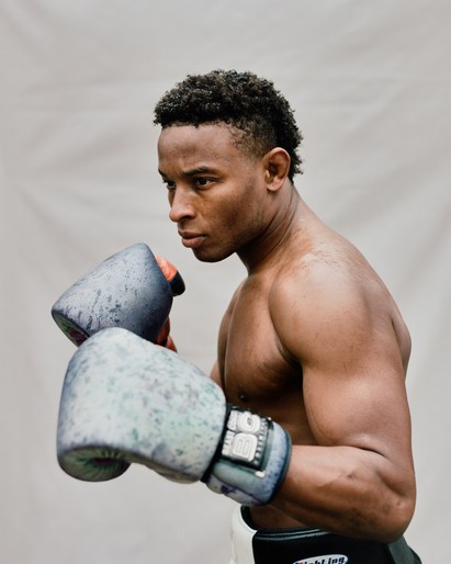 Muscular male boxer with dark hair and fair skin wearing worn boxing gloves in a close-up studio shot. Dark belt visible at waist.