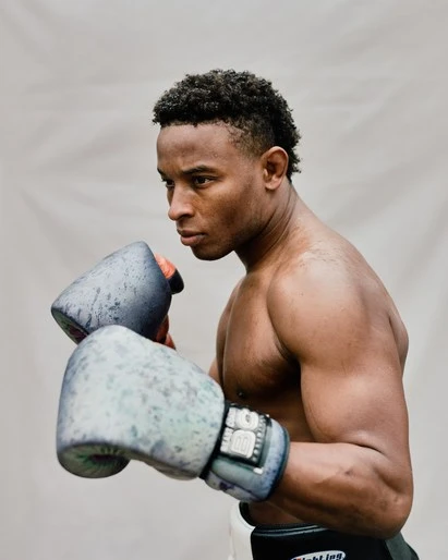 Muscular male boxer with dark hair and fair skin wearing worn boxing gloves in a close-up studio shot. Dark belt visible at waist.
