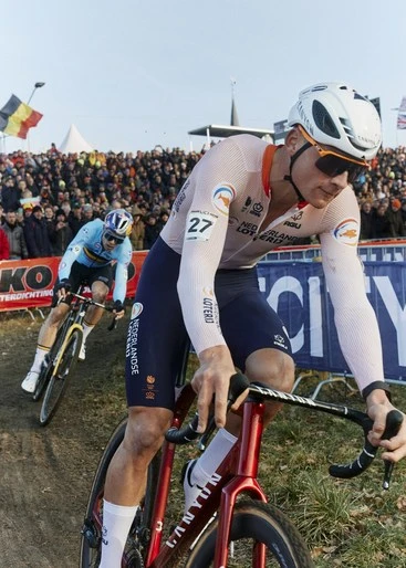 Cyclist in action during a race, wearing a helmet and team gear, with a crowd in the background and racing bike in focus.