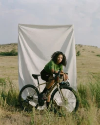 Woman leaning on a bicycle in front of a white fabric backdrop set in a grassy outdoor landscape