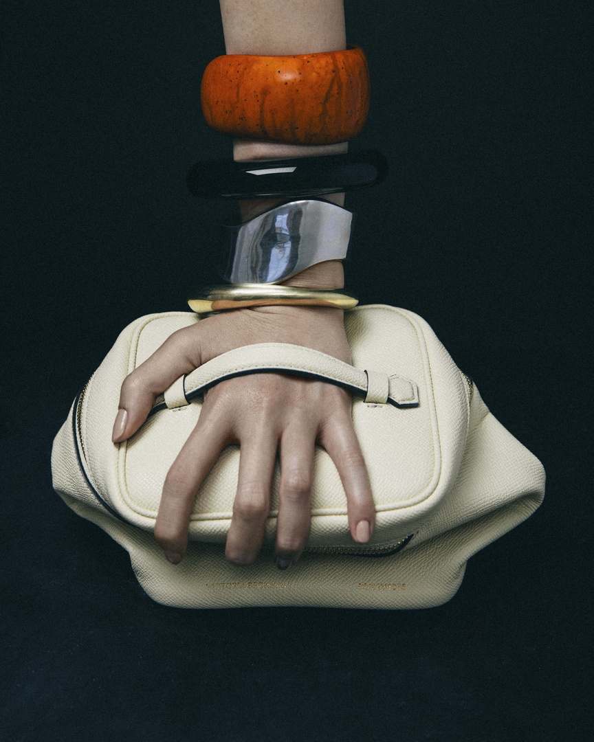 An arm with stacked bracelets holds a light beige purse against a dark background in a fashion-forward studio product shot.